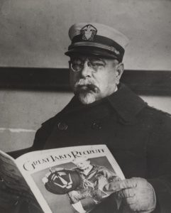 Photograph showing John Philip Sousa, with a white beard and smoking a cigar, reading the "Great Lakes Naval Recruit" magazine.
