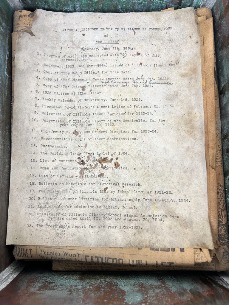 List of the contents of the University Library time capsule, found inside the copper box in the cornerstone. The paper is moldy and scraps of disintegrated paper can be seen on top. The edges of the copper box are visibly oxidized.